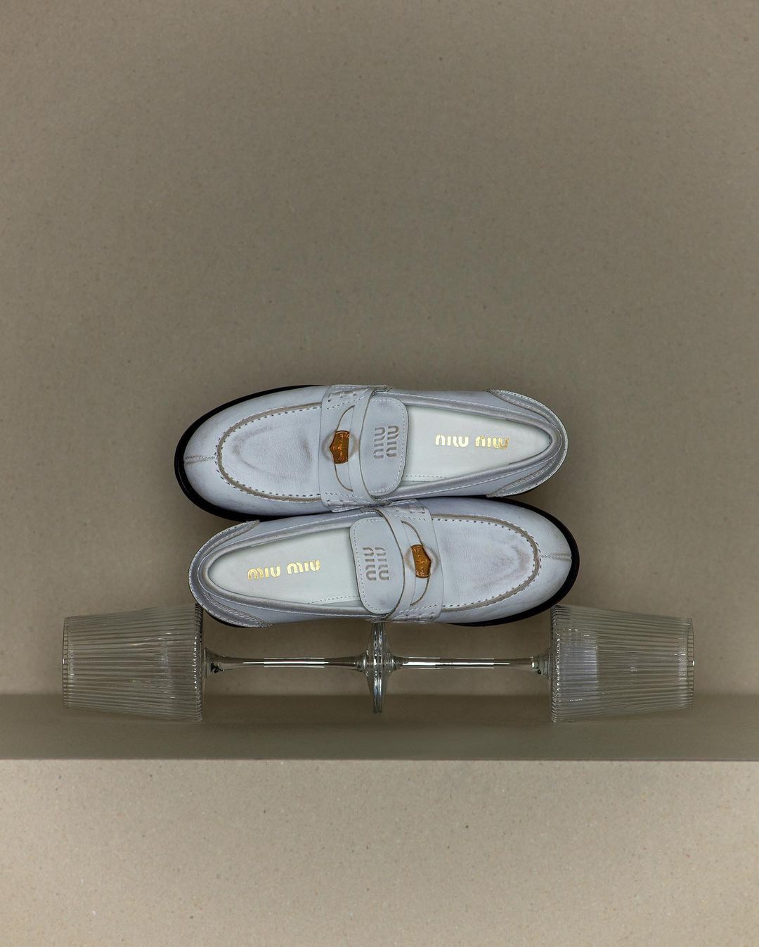 Two pairs of Miu Miu white leather loafers stacked on each other, displayed on a light brown surface with a glass object.