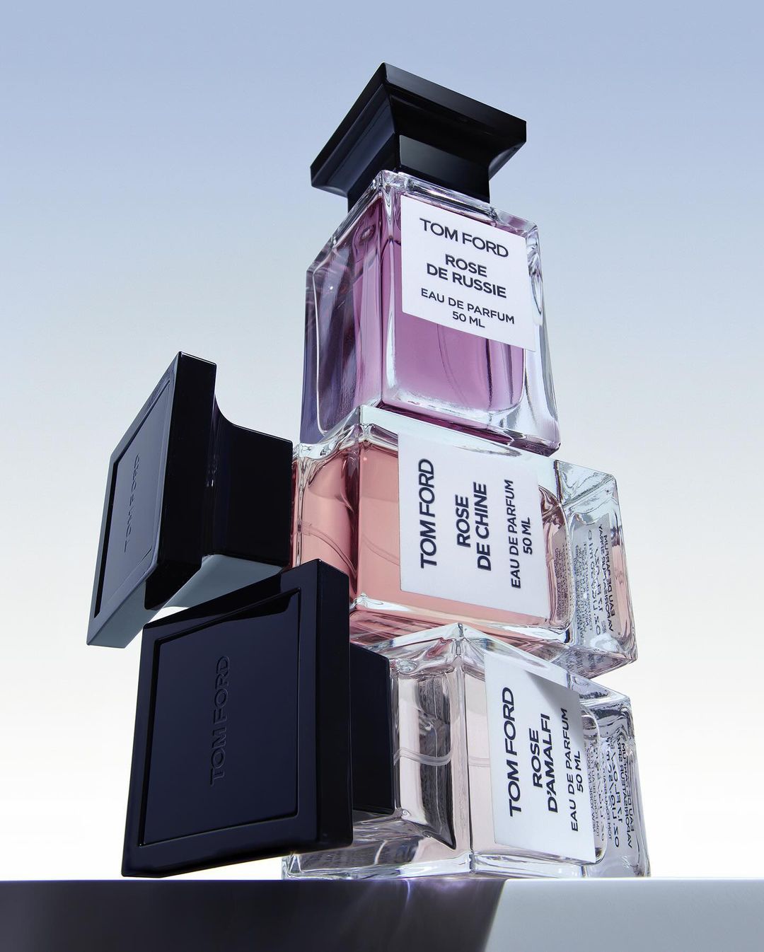 Stacked Tom Ford fragrance bottles with black caps against a pale gradient background