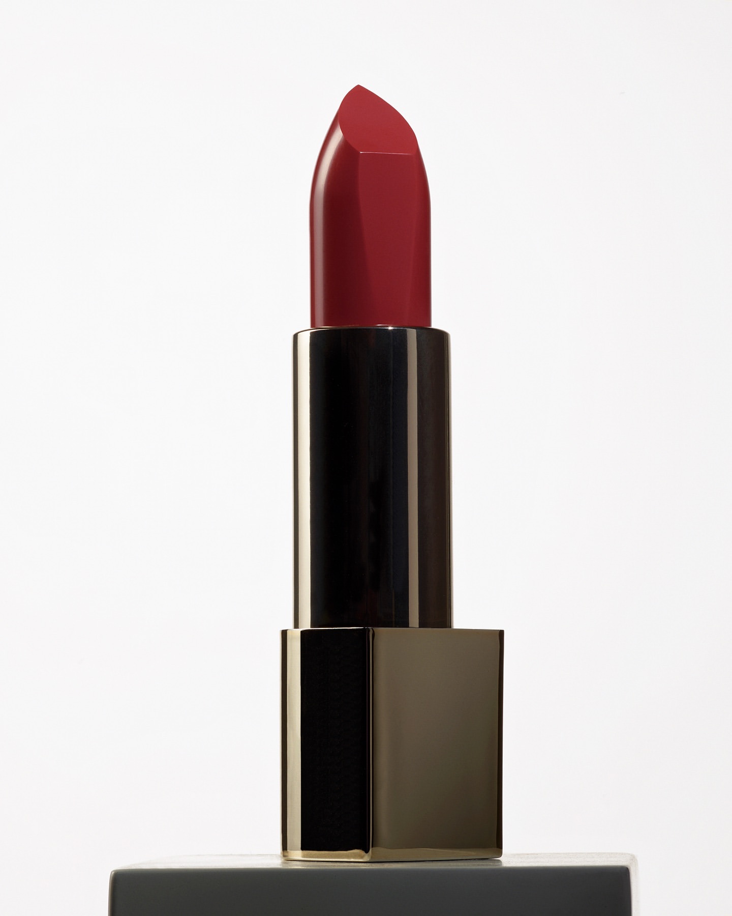 Close-up studio shot of a dark red lipstick with a black and gold tube, reflecting light against a solid white background.