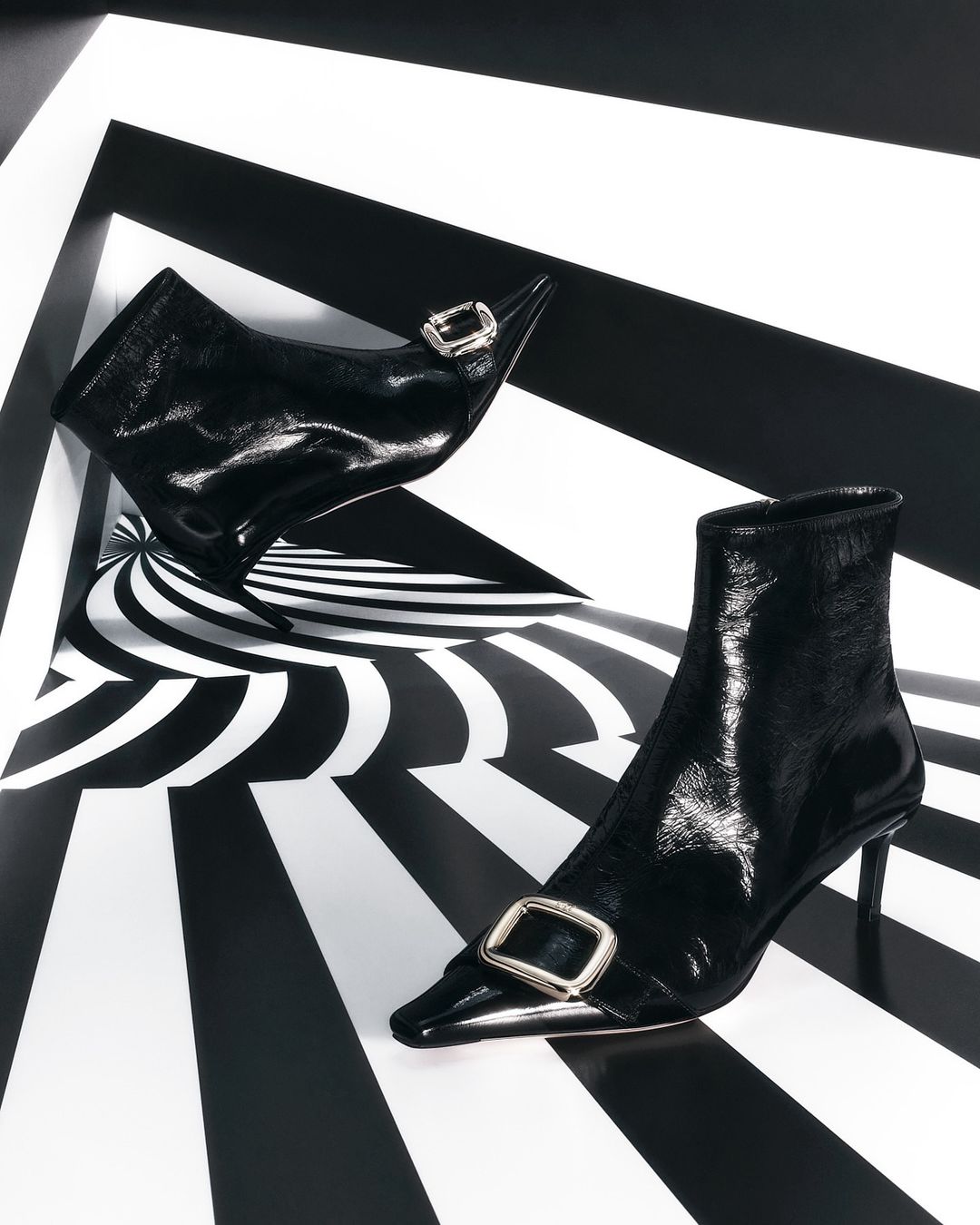 Elegant black patent leather ankle boots with decorative buckles, set against a geometric black and white background.