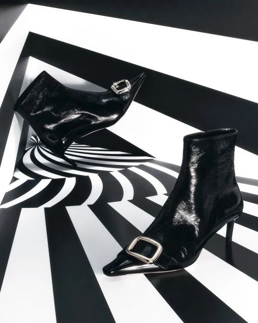 Elegant black patent leather ankle boots with decorative buckles, set against a geometric black and white background.
