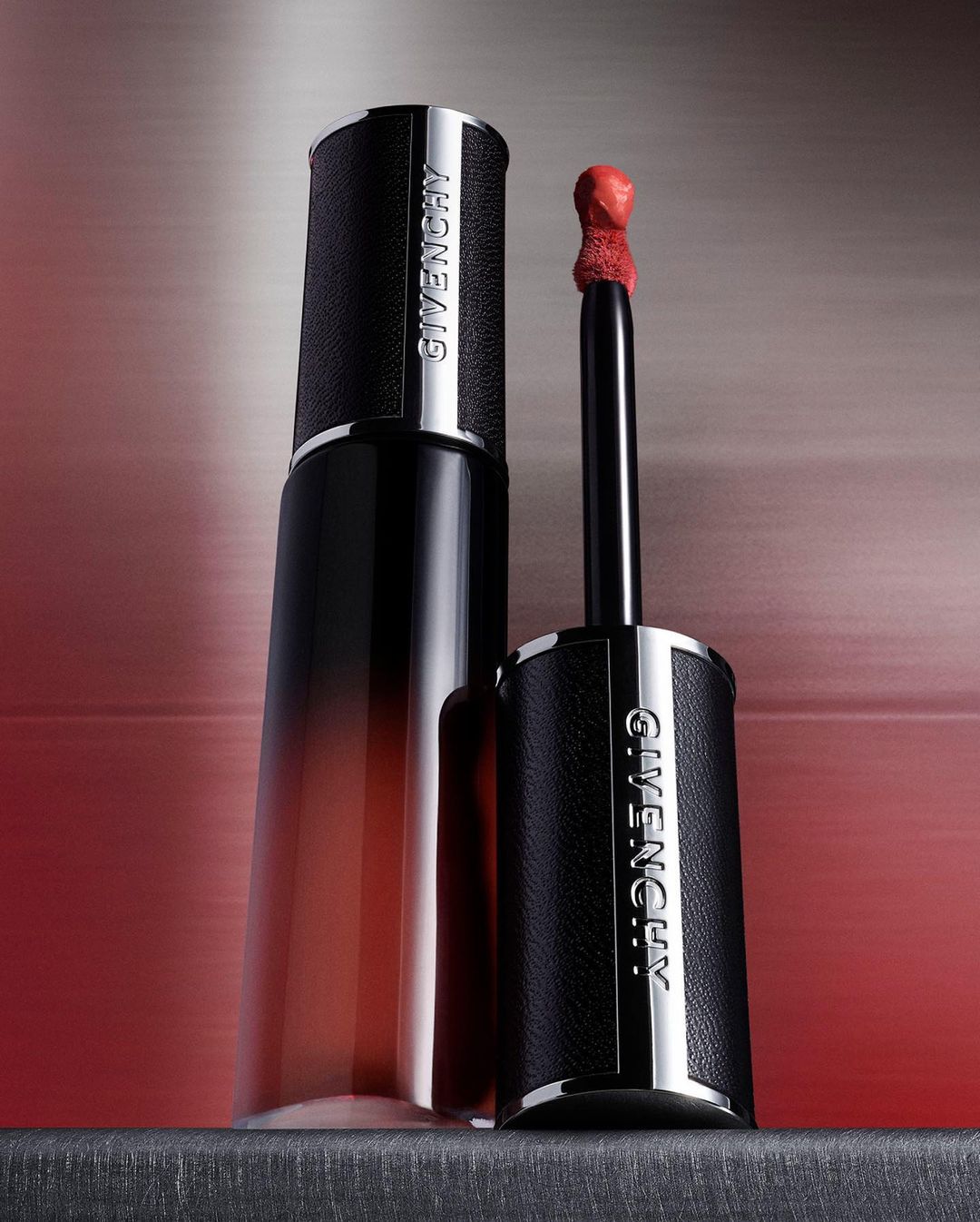 Givenchy Le Rouge Interdit Cream Velvet lipstick with a black leather tube, silver accents, and a red-colored applicator, set against a blurred red backdrop.