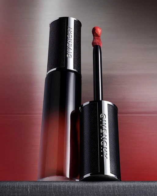 Givenchy Le Rouge Interdit Cream Velvet lipstick with a black leather tube, silver accents, and a red-colored applicator, set against a blurred red backdrop.