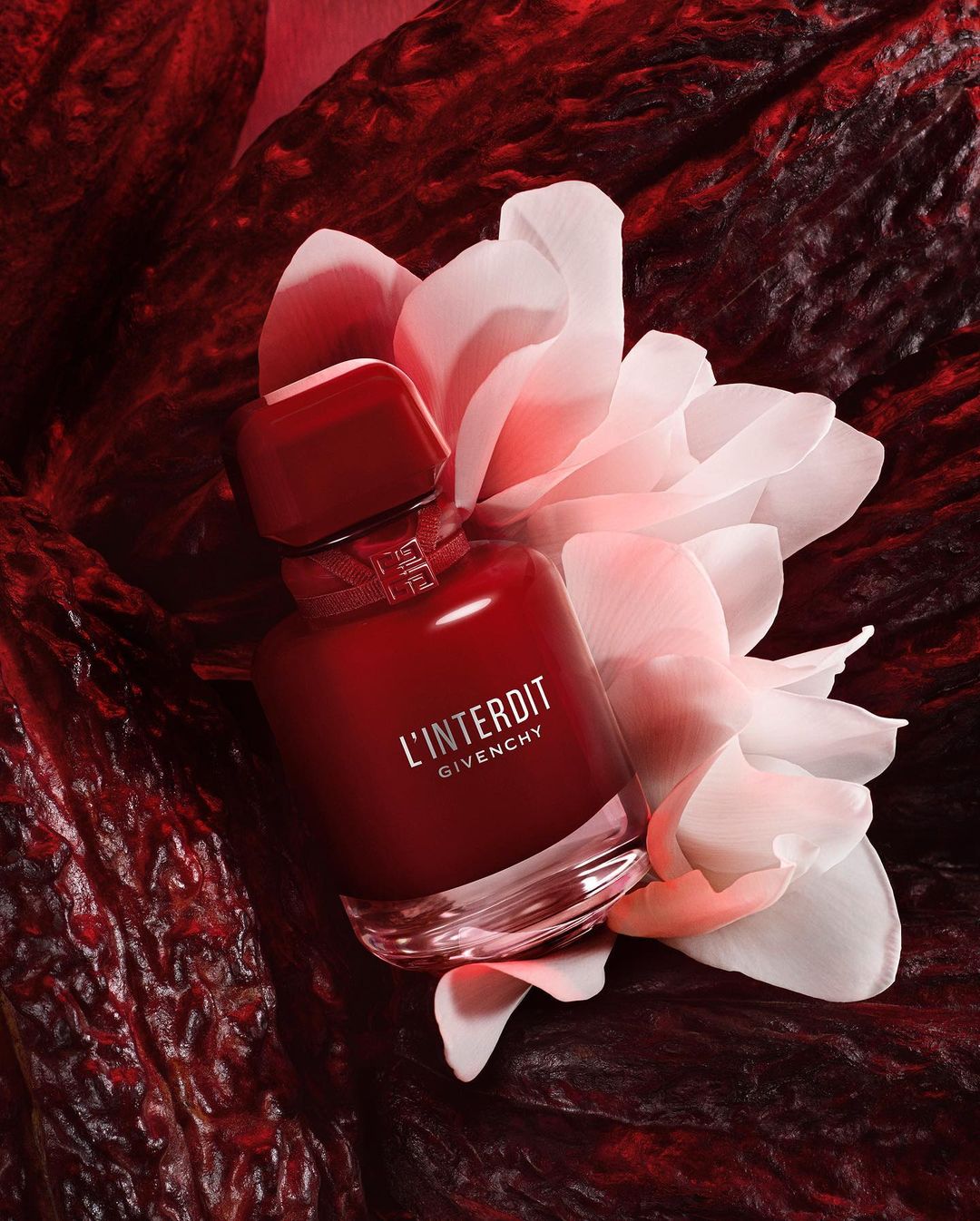 Studio shot of a red perfume bottle with white flowers in a dark red textured background; the perfume is L'Interdit by Givenchy.