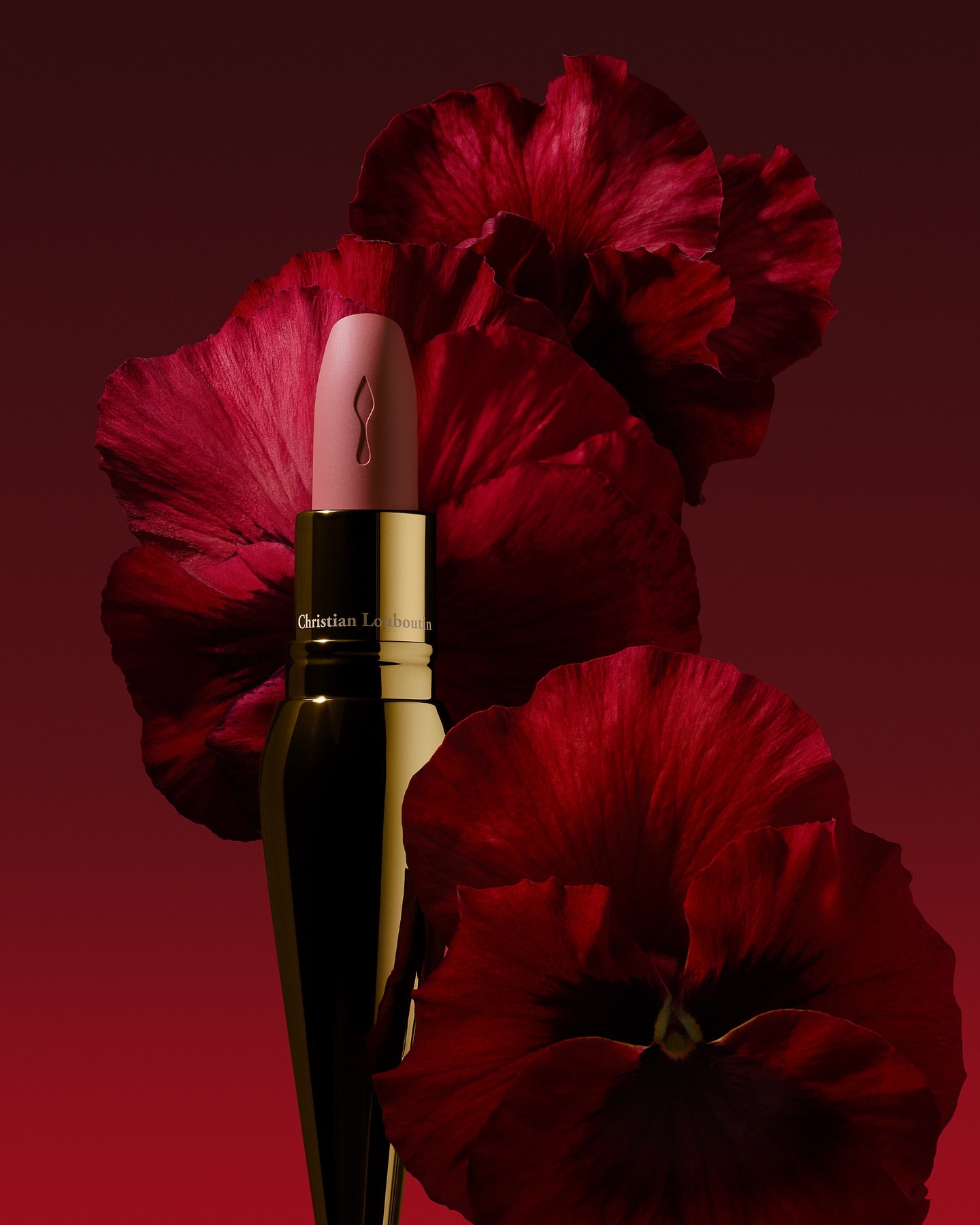 Close-up studio shot of light pink Christian Louboutin lipstick tube with a gold base against dark red hibiscus flowers.