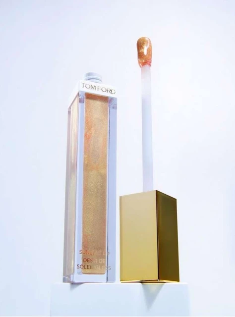 Tom Ford Soleil lip gloss in gold packaging on a white surface, featuring a long applicator and golden hues.