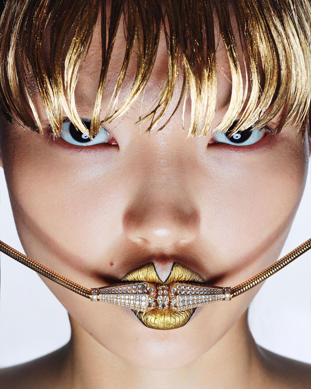Close-up portrait of a stylish adult female with gold hair and creative gold and diamond lip jewelry against a bright background.