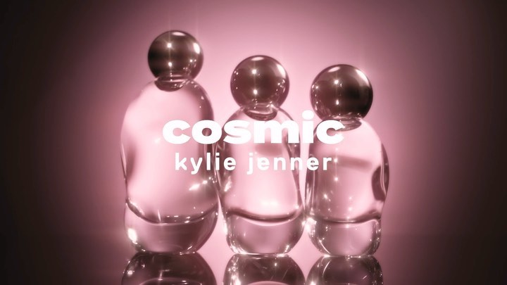 Three clear glass perfume bottles with spherical tops sit in a row, lit with pink and displaying 'cosmic kylie jenner' text overlay.