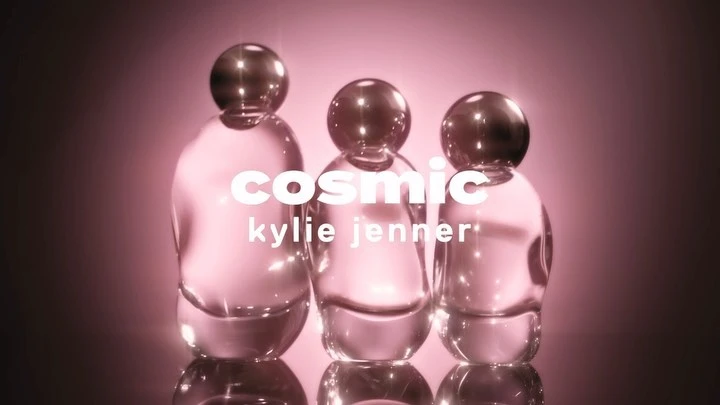 Three clear glass perfume bottles with spherical tops sit in a row, lit with pink and displaying 'cosmic kylie jenner' text overlay.