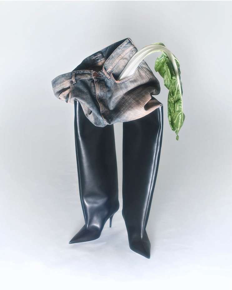 Fashion editorial: Jeans and black thigh-high stiletto boots on a white background, with a piece of green vegetable.