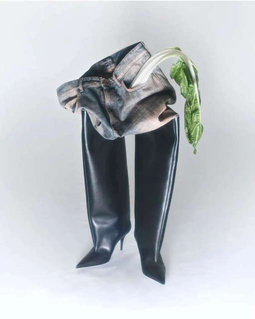 Fashion editorial: Jeans and black thigh-high stiletto boots on a white background, with a piece of green vegetable.