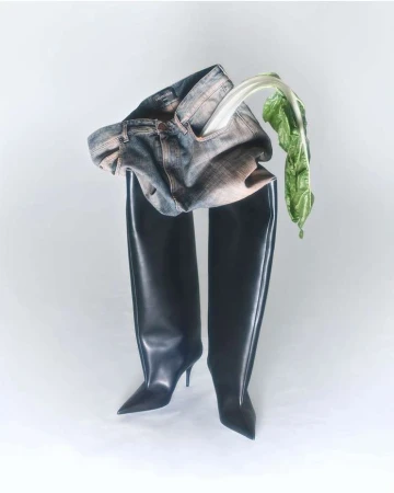 Fashion editorial: Jeans and black thigh-high stiletto boots on a white background, with a piece of green vegetable.