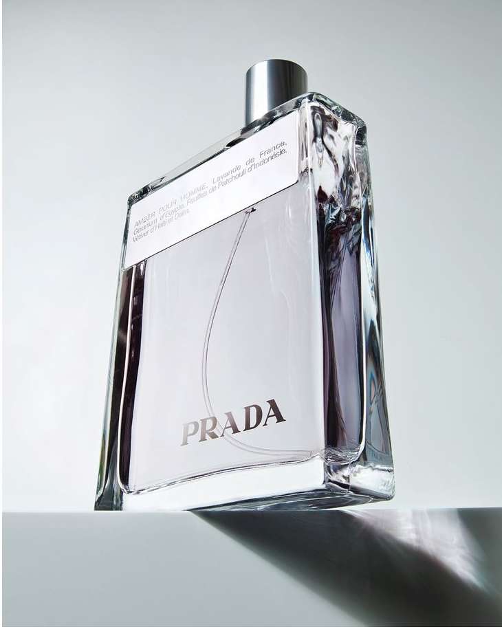 Product shot of a rectangular glass bottle of Prada fragrance with a silver cap, against a neutral white background with soft lighting.
