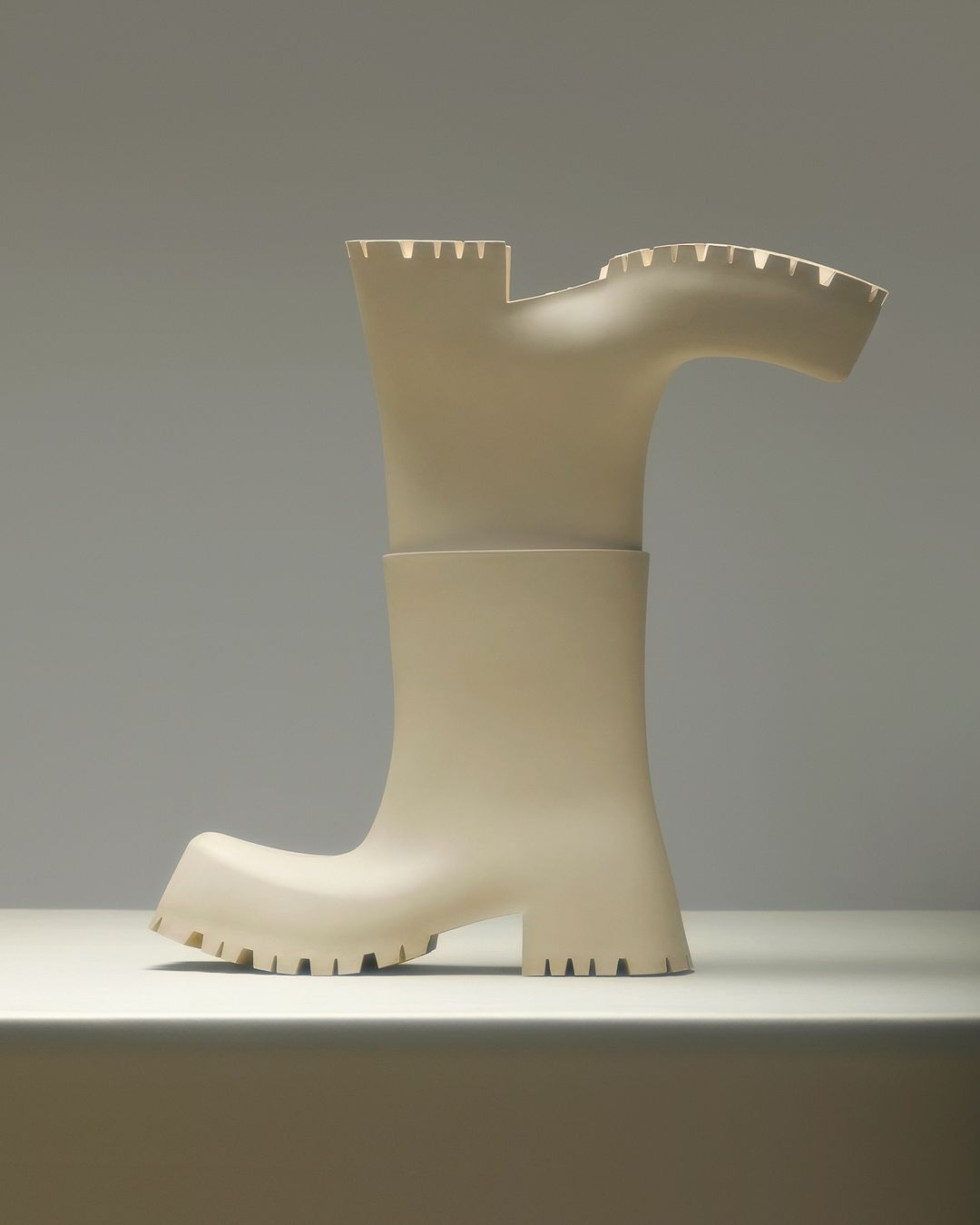 Abstract boot sculpture made of matte off-white material displayed on a white surface with a neutral-tone background in a studio setting.