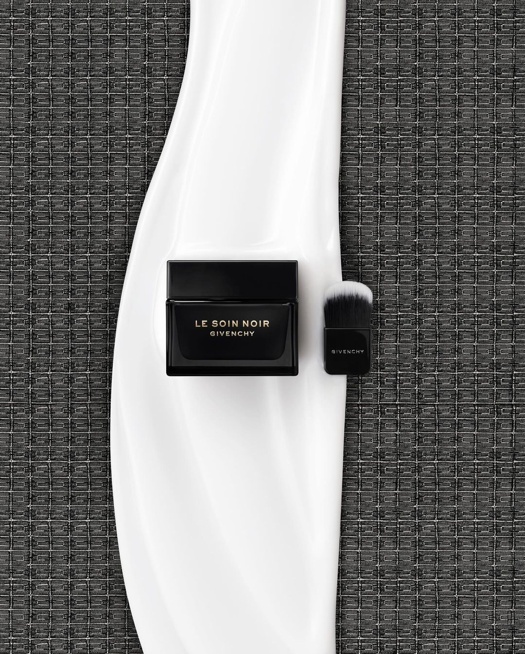 High-angle shot of Le Soin Noir Givenchy cream jar and brush on a glossy white surface against a textured grey background.