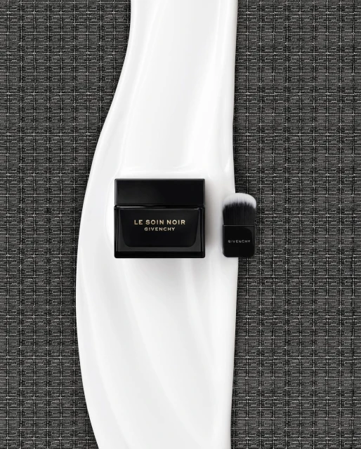 High-angle shot of Le Soin Noir Givenchy cream jar and brush on a glossy white surface against a textured grey background.