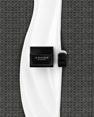 High-angle shot of Le Soin Noir Givenchy cream jar and brush on a glossy white surface against a textured grey background.