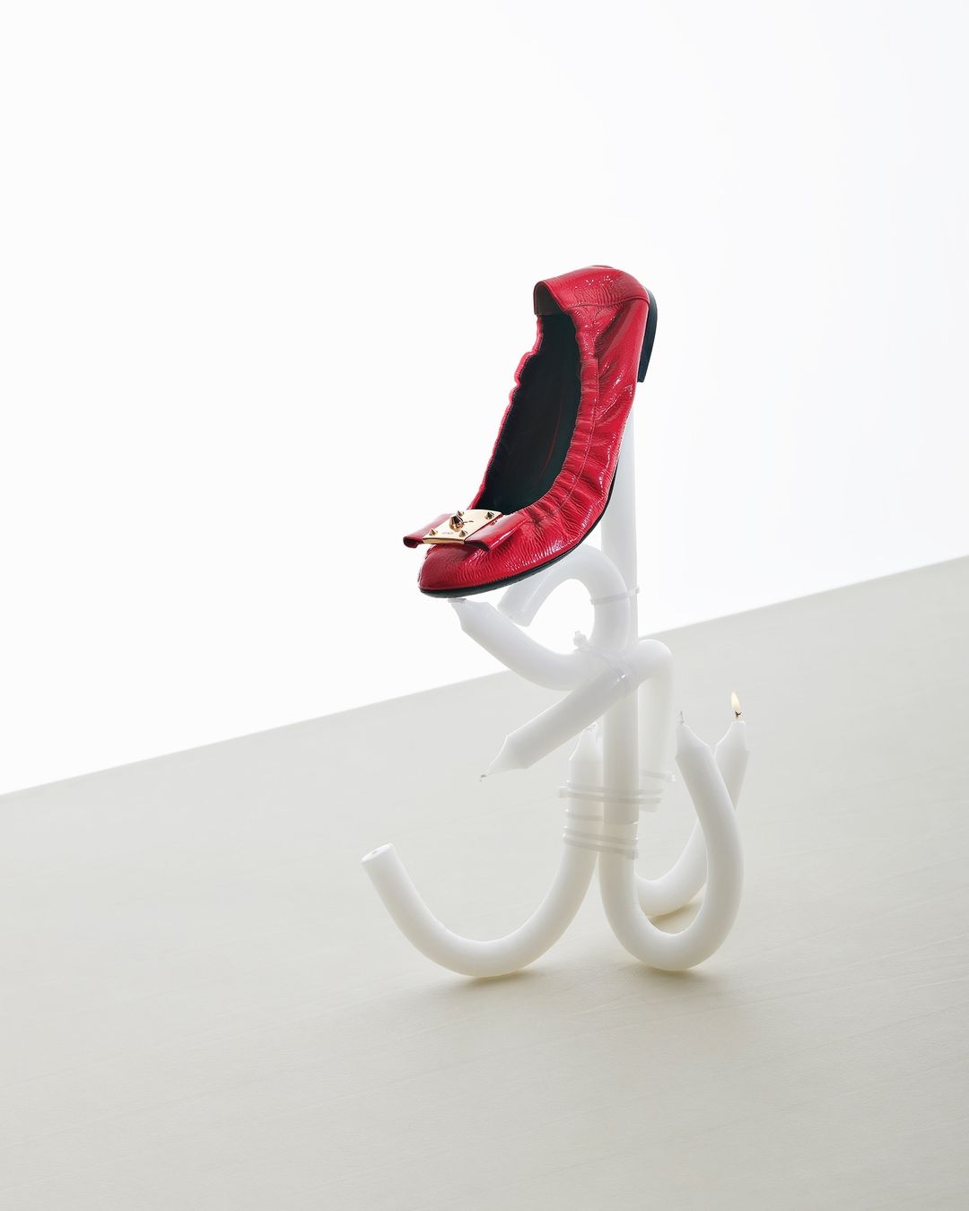 Studio shot of a single red ballet flat with a gold buckle on a white candle stand against a white backdrop.