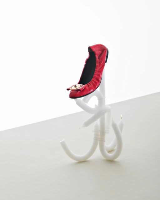 Studio shot of a single red ballet flat with a gold buckle on a white candle stand against a white backdrop.