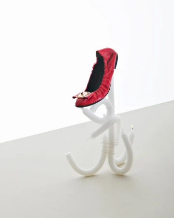 Studio shot of a single red ballet flat with a gold buckle on a white candle stand against a white backdrop.