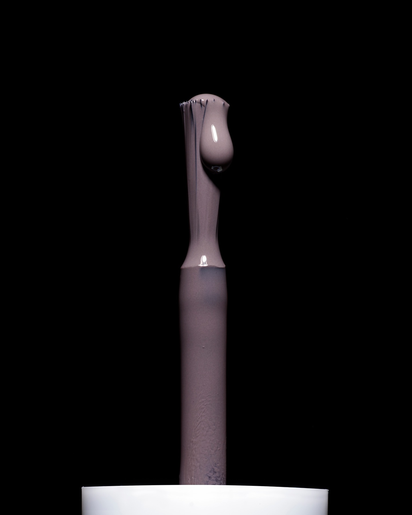 Close-up of a nail polish brush applicator with a droplet of mauve-colored nail polish against a dark black studio background.