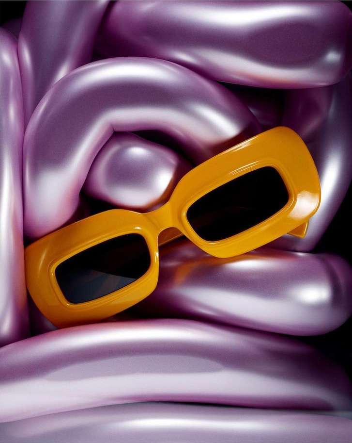 A pair of yellow, oversized sunglasses is the primary focus against a backdrop of shiny, curved lilac forms. Product shot.