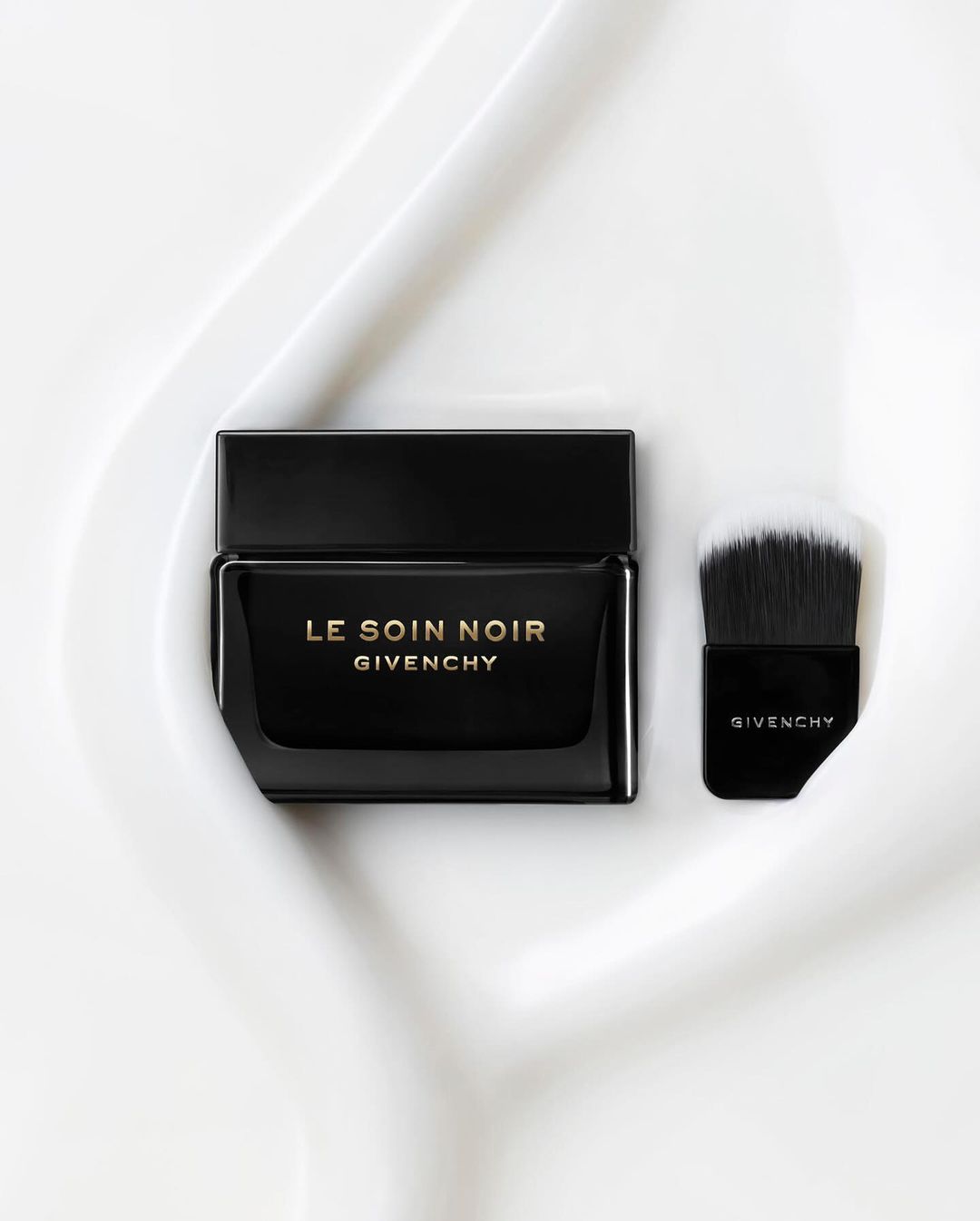 Elegant black Givenchy Le Soin Noir cream jar and applicator brush sit on a textured white surface. Beauty and luxury product shot.