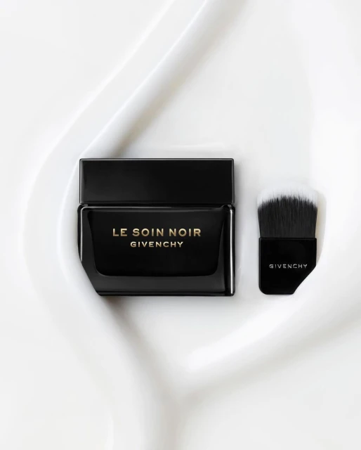 Elegant black Givenchy Le Soin Noir cream jar and applicator brush sit on a textured white surface. Beauty and luxury product shot.