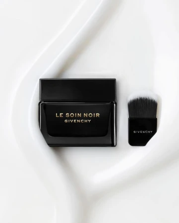 Elegant black Givenchy Le Soin Noir cream jar and applicator brush sit on a textured white surface. Beauty and luxury product shot.