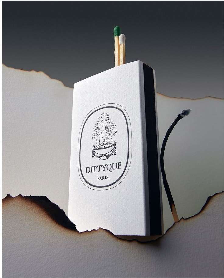 Close-up studio shot of a Diptyque matchbox with matches, alongside a burnt piece of paper, set against a dark gray gradient backdrop.