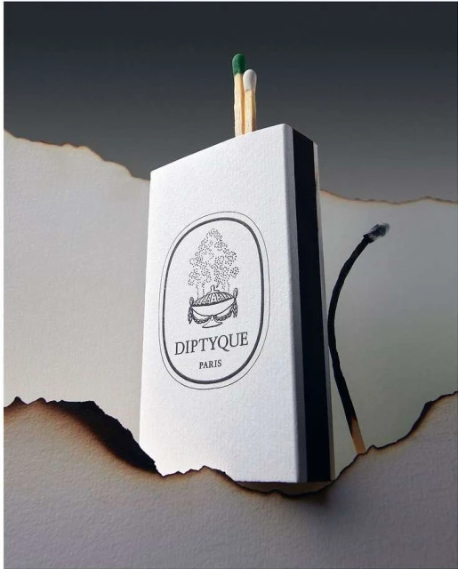 Close-up studio shot of a Diptyque matchbox with matches, alongside a burnt piece of paper, set against a dark gray gradient backdrop.