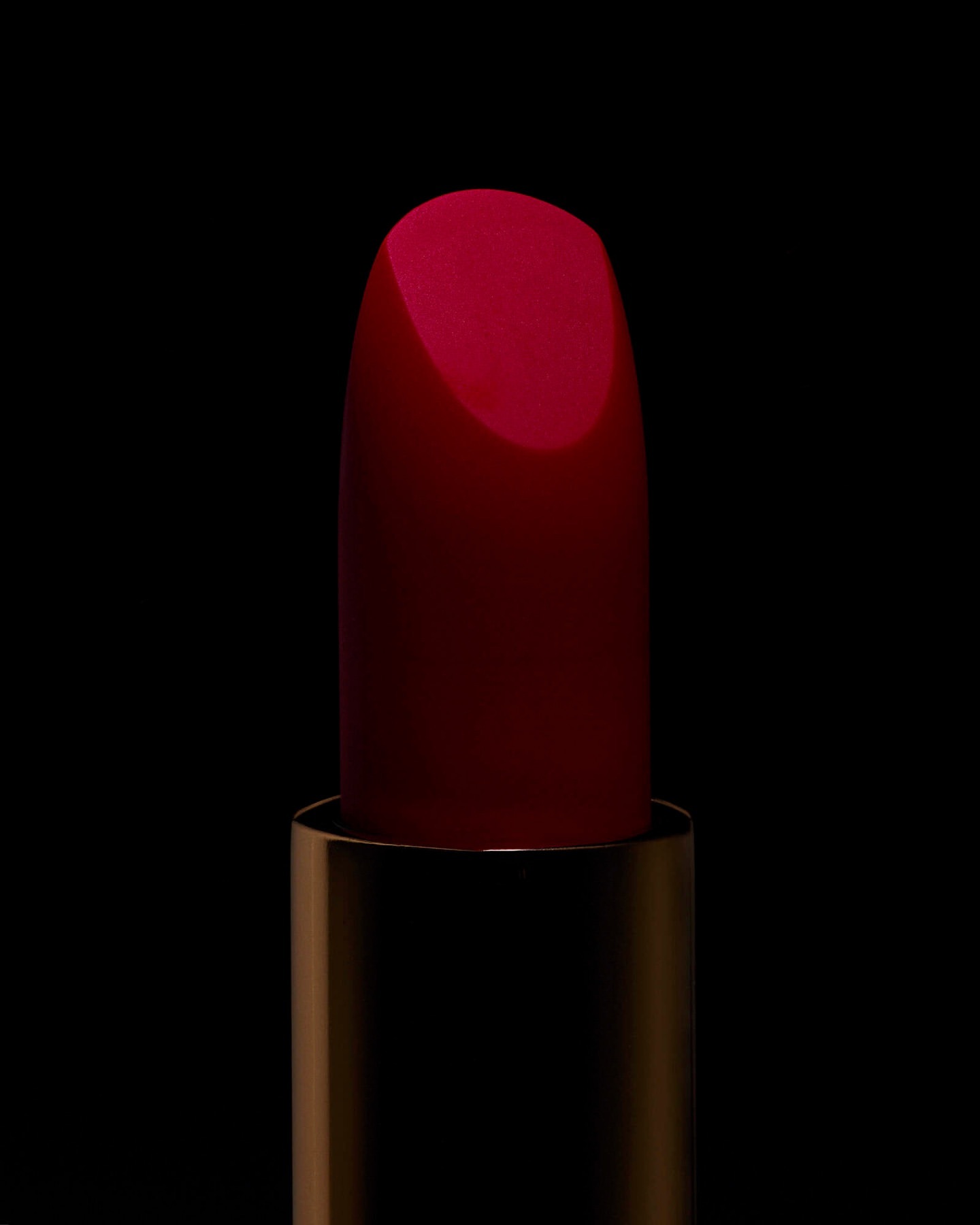 Extreme macro close-up of a crimson red lipstick bullet emerging from its metallic golden case against a pitch-black background.