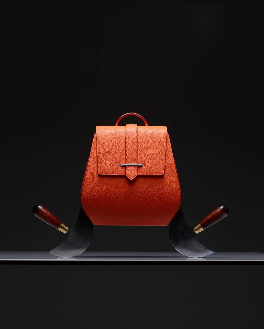 Orange leather backpack on a shelf with cleavers on either side against a black background, showcasing a blend of fashion and edge.
