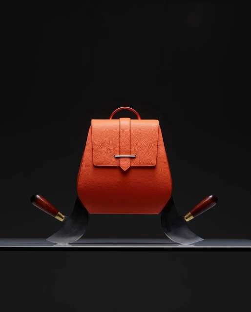 Orange leather backpack on a shelf with cleavers on either side against a black background, showcasing a blend of fashion and edge.