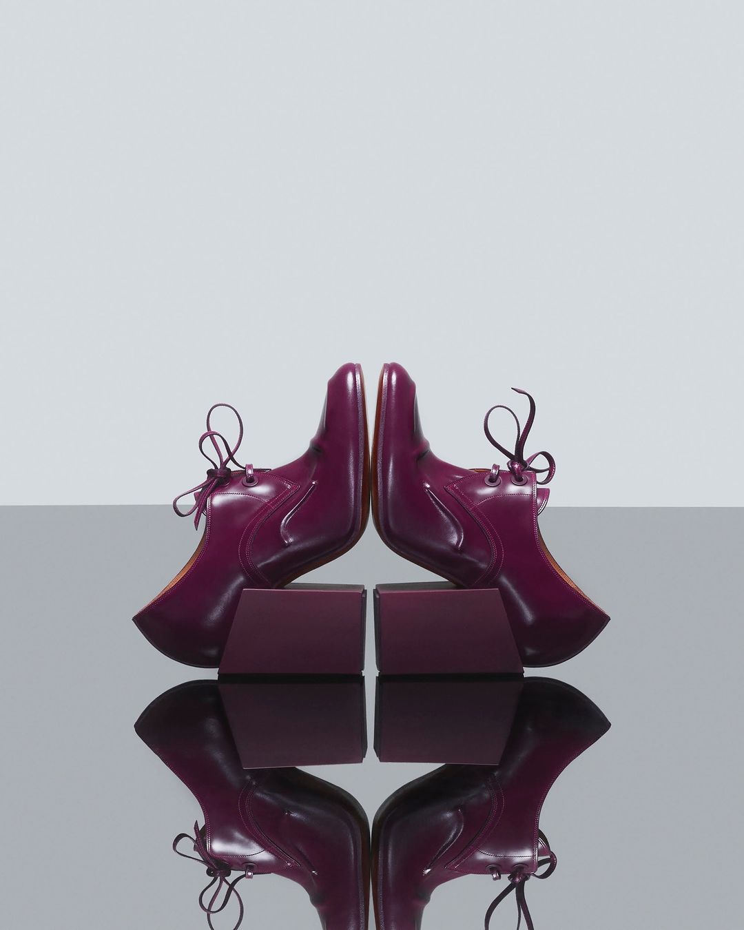 Symmetrical product shot of two plum-colored, patent-leather lace-up shoes resting on matching blocks against a light-grey background.