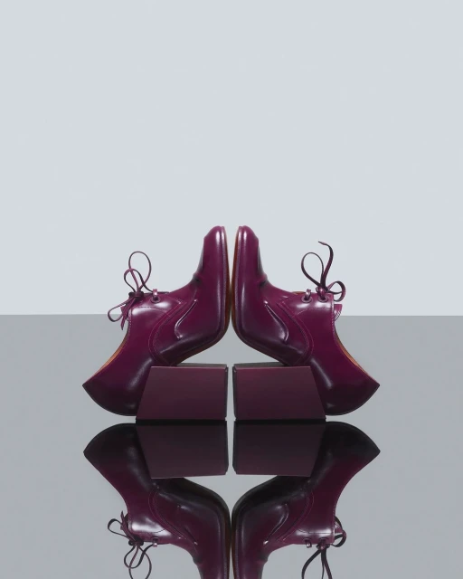 Symmetrical product shot of two plum-colored, patent-leather lace-up shoes resting on matching blocks against a light-grey background.