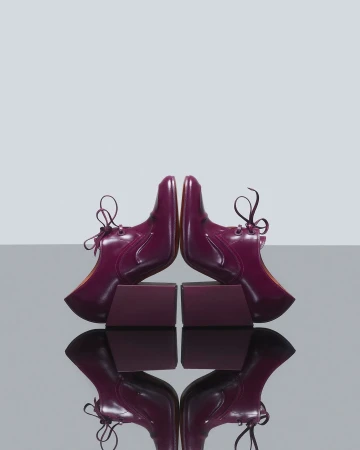 Symmetrical product shot of two plum-colored, patent-leather lace-up shoes resting on matching blocks against a light-grey background.