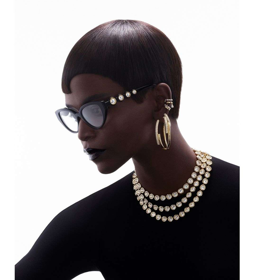 Close-up portrait of a Black woman wearing cat-eye glasses, layered crystal necklaces, and hoop earrings against a white background.