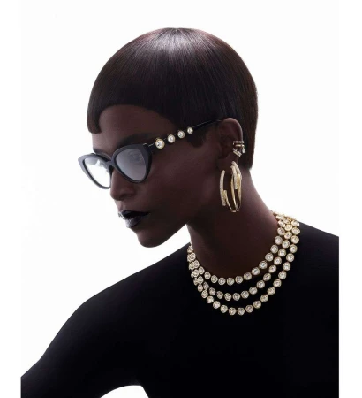 Close-up portrait of a Black woman wearing cat-eye glasses, layered crystal necklaces, and hoop earrings against a white background.