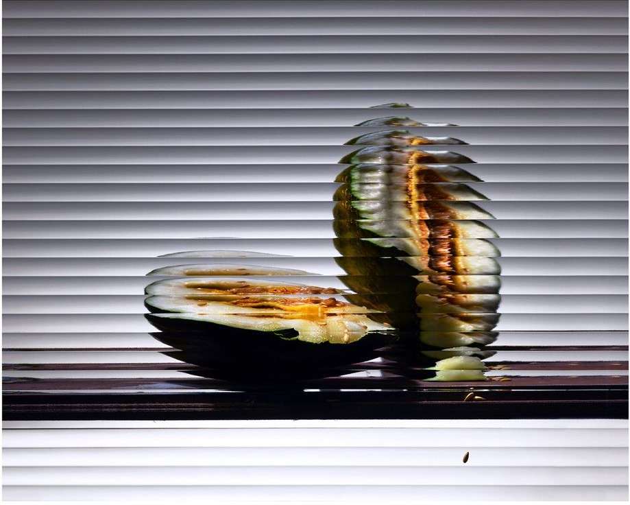 Sliced melon on a dark surface, set against horizontal window blinds. The melon's vibrant interior contrasts with the neutral blinds.