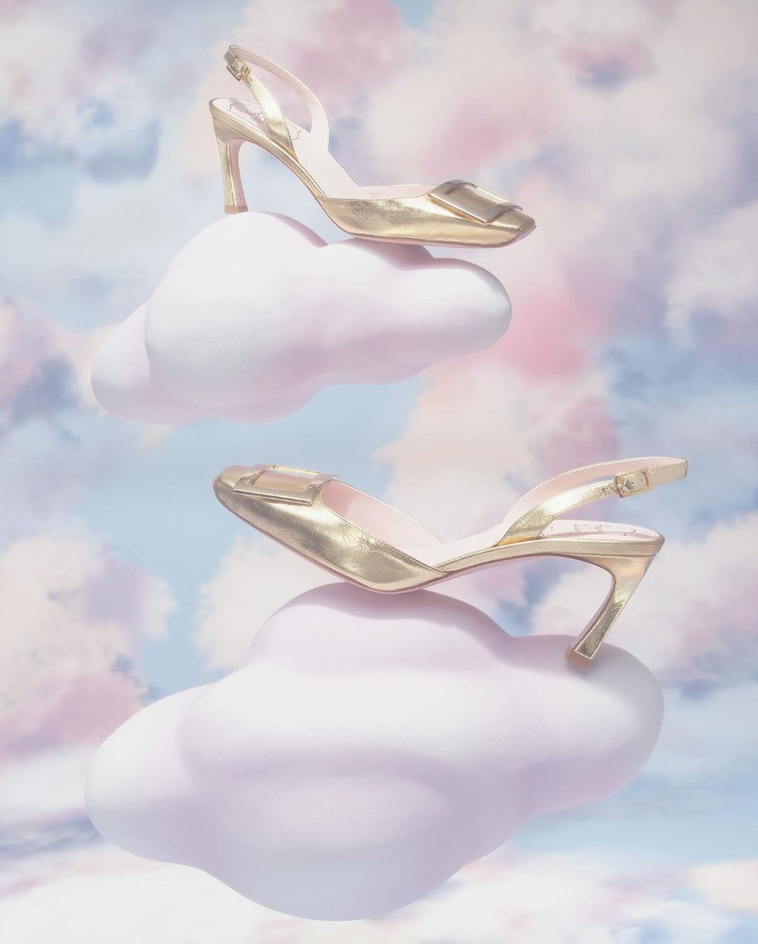 Elevated fashion shot of two gold slingback high heels perched on fluffy pink clouds against a soft pastel blue and pink sky backdrop.