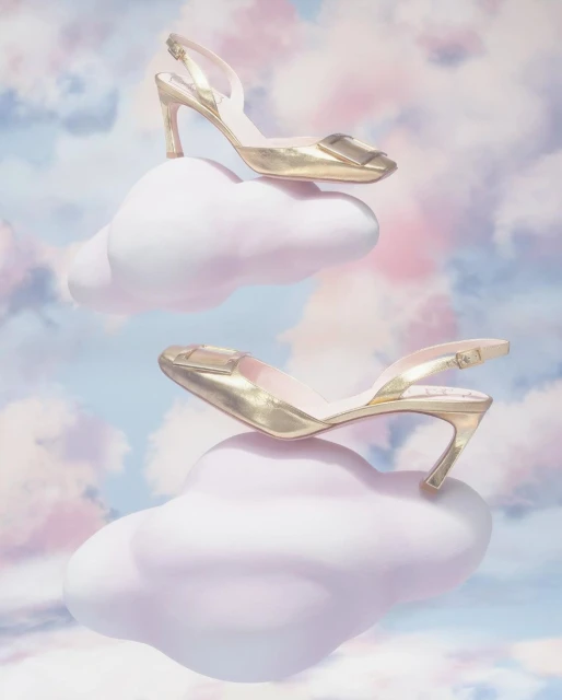 Elevated fashion shot of two gold slingback high heels perched on fluffy pink clouds against a soft pastel blue and pink sky backdrop.