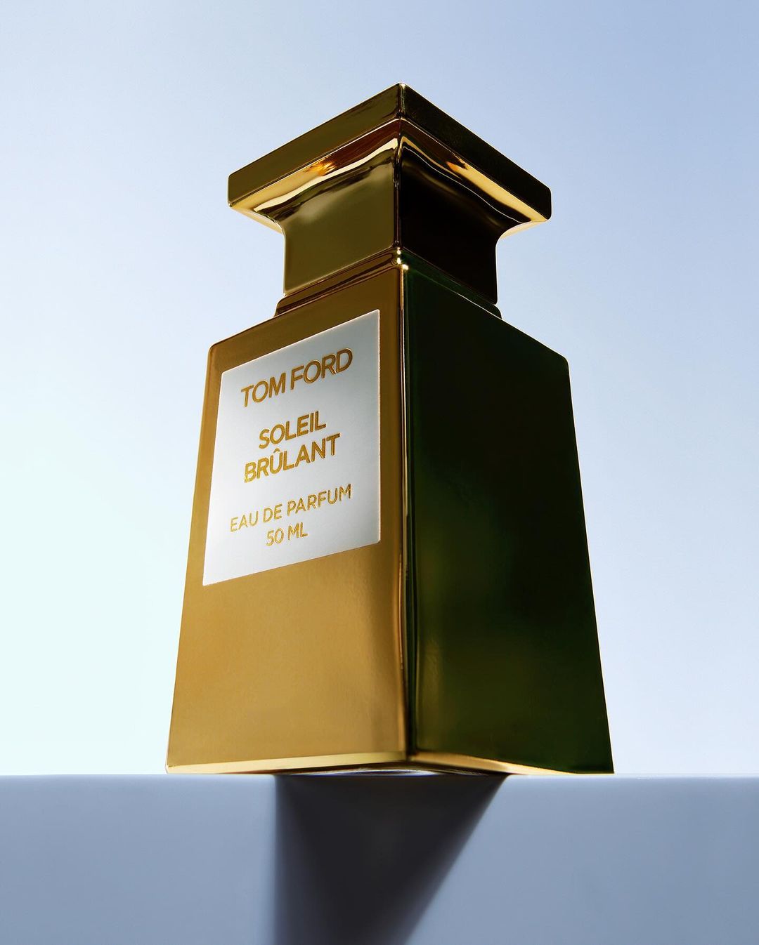 Tom Ford Soleil Brulant perfume bottle in gold and silver standing upright against a pale blue sky.