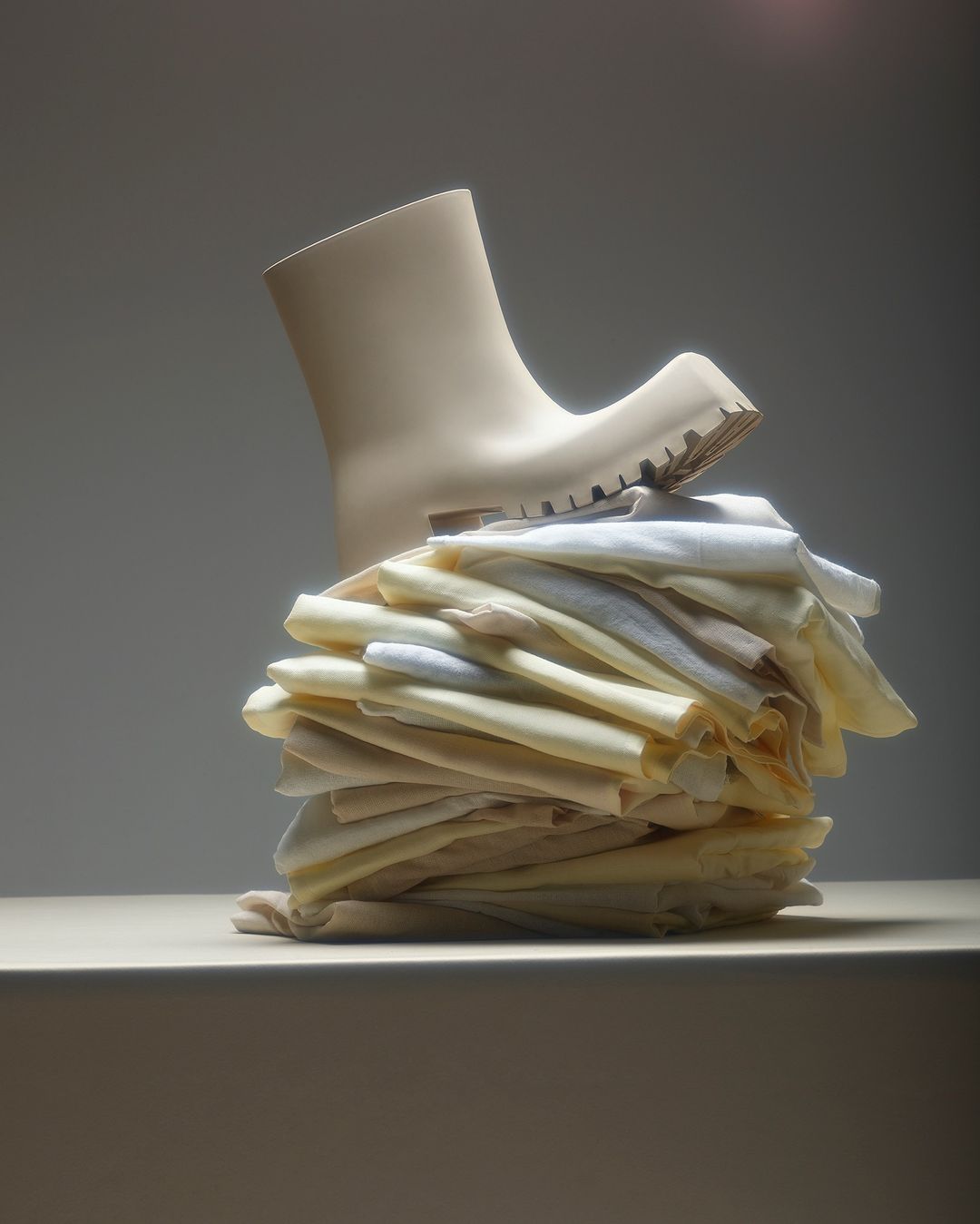 Cream colored rubber boot sits atop a stack of folded white cotton t-shirts on a cream surface against a pale studio backdrop.