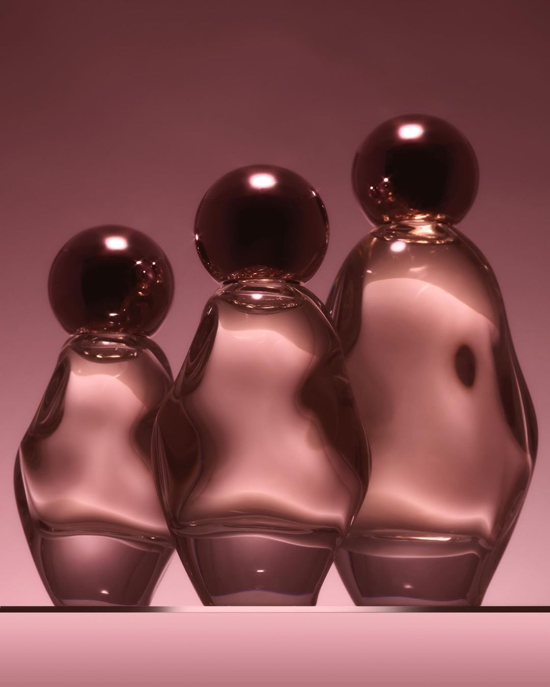 Three glass perfume bottles in varying sizes arranged on a pink surface against a pink backdrop, with light reflecting on their surfaces.