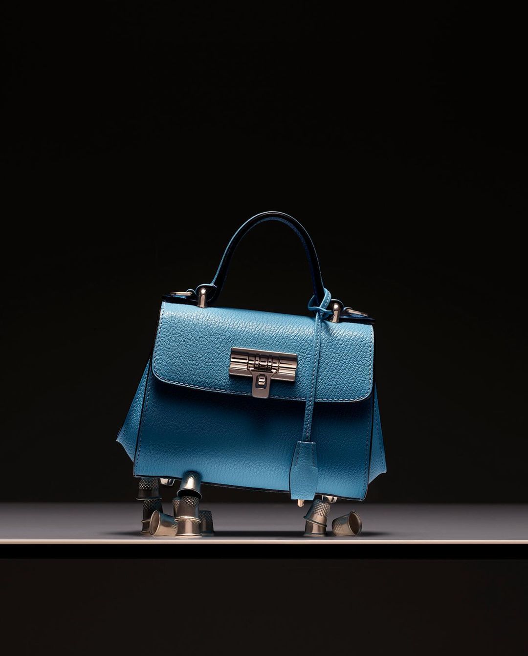 Stylish blue leather purse with a silver clasp and metal legs, set on a gray shelf against a dark background, fashion accessory.