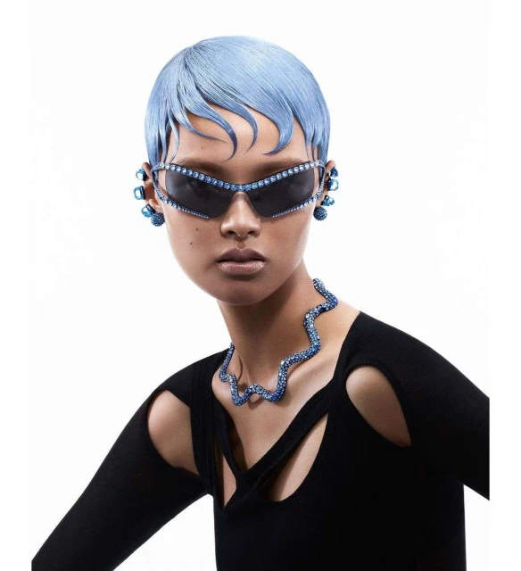 A woman with light blue hair wearing sunglasses and a necklace with blue gems, posing in front of a white background in a fashion style.