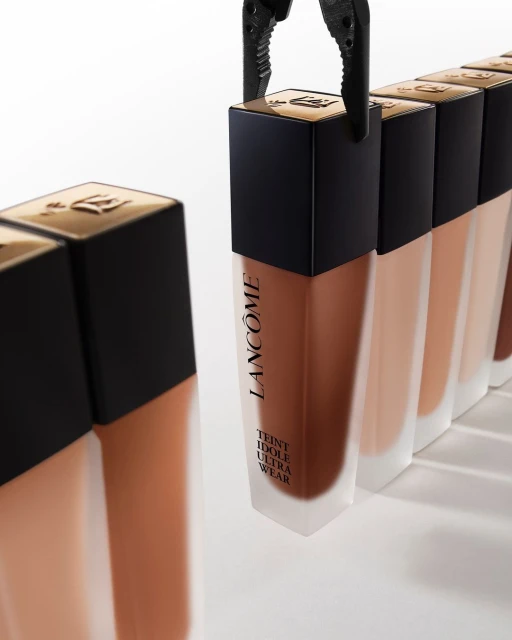 Lancôme Teint Idole Ultra Wear foundation bottles in varying shades of tan, held by black tweezers against a white backdrop.