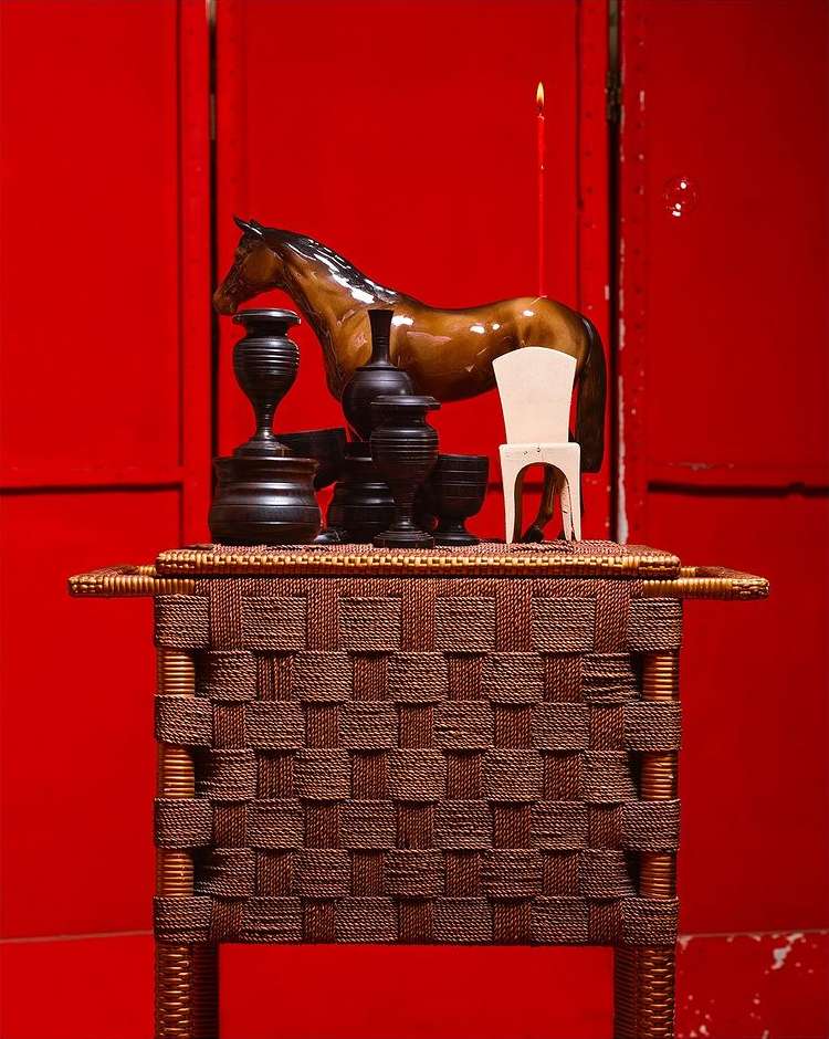 Still life featuring a brown horse figurine, small white chair, black vases and candle on a woven table against a red backdrop.