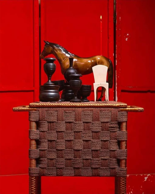 Still life featuring a brown horse figurine, small white chair, black vases and candle on a woven table against a red backdrop.